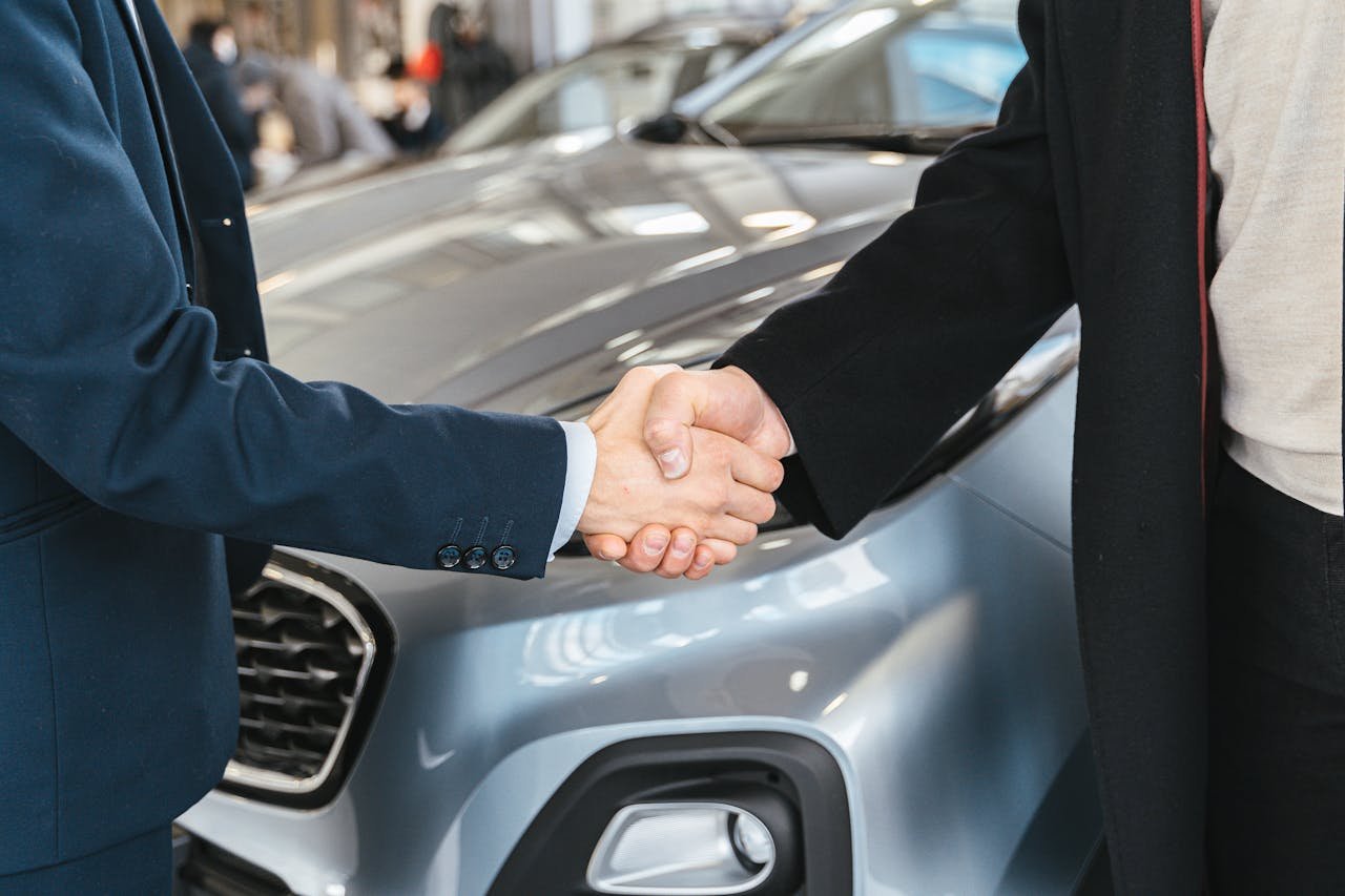 why-choose-us A close-up of two people shaking hands in front of a car in an indoor setting.